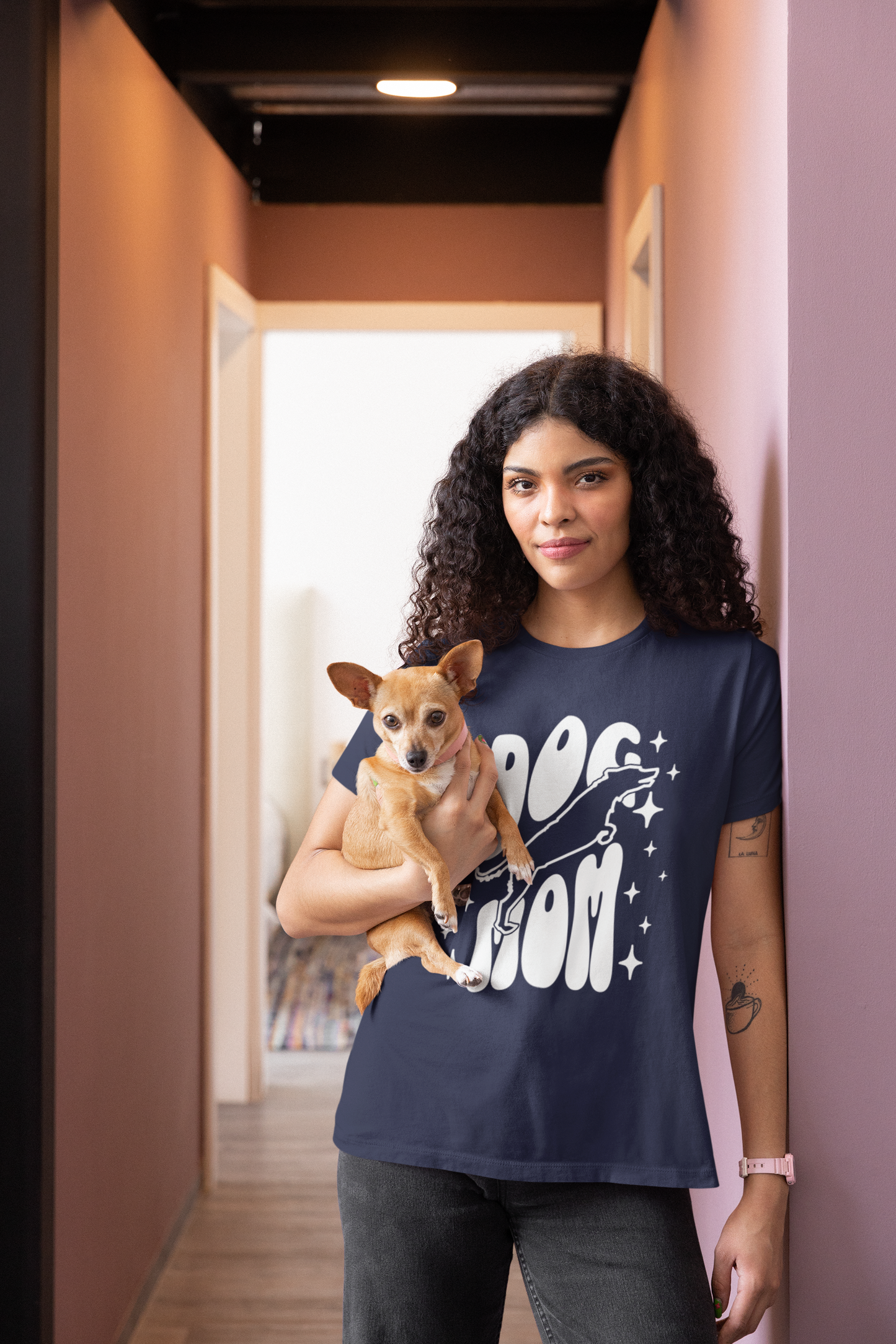 Woman wearing a navy blue t-shirt with 'Dog Mom' text, holding a small dog in a hallway.